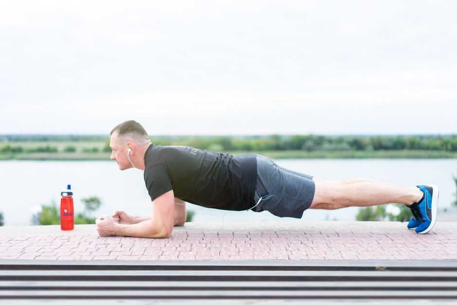 Man in sportswear doing a plank exercise outdoors near a lake with headphones on.